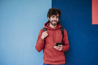 Young smiling man with smart phone near colorful wall - EBBF07146