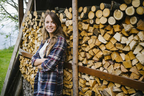 Lächelnde junge Frau mit verschränkten Armen vor Baumstämmen stehend - GIOF15639