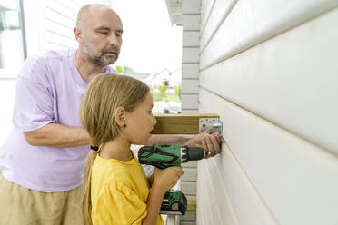 Vater lehrt Tochter mit Schraubenzieher in der Nähe der Holzwand am Haus zu arbeiten - KMKF01893
