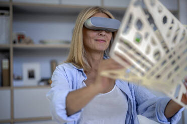 Smiling mature woman with VR glasses holding 3d print at home - RIBF01213