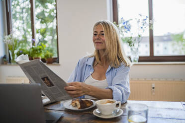 Smiling mature woman reading newspaper with food at table - RIBF01220