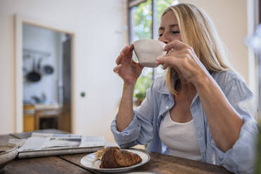 Mature woman drinking coffee with croissant on table at home - RIBF01223
