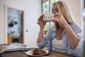 Reife Frau trinkt Kaffee mit Croissant auf dem Tisch zu Hause - RIBF01223