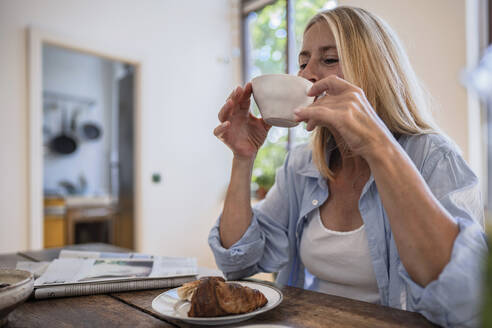 Reife Frau trinkt Kaffee mit Croissant auf dem Tisch zu Hause - RIBF01223