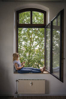 Kontemplative reife Frau, die zu Hause auf der Fensterbank sitzt - RIBF01232
