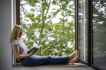 Woman reading book sitting on window sill at home - RIBF01233