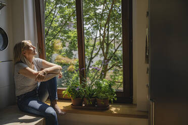 Mature woman enjoying sunlight sitting by window in kitchen - RIBF01243