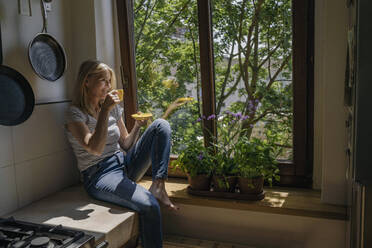 Mature woman drinking coffee sitting on window sill at home - RIBF01247