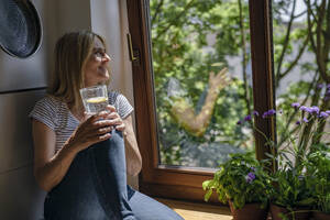 Reife Frau träumt mit einem Glas Wasser am Fenster - RIBF01249