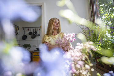 Lächelnde reife Frau schaut durch das Fenster zu Hause - RIBF01309