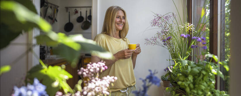 Glückliche reife Frau, die eine Tasse Kaffee in der Nähe des Fensters zu Hause steht - RIBF01312