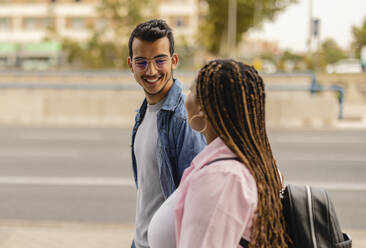 Smiling young man walking with girlfriend at footpath - JCCMF08043