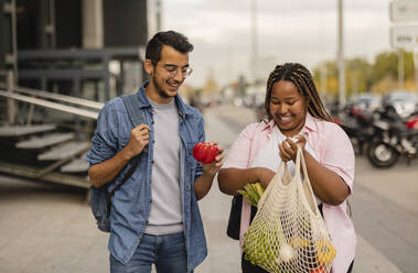 Fröhlicher junger Mann mit Frau, die einen Sack mit Gemüse auf dem Fußweg hält - JCCMF08044
