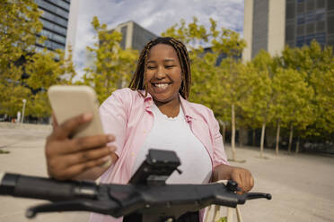 Happy curvy woman with electric push scooter using smart phone - JCCMF08051