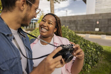 Fröhliche junge Frau steht mit einem Mann, der eine Kamera hält, im Park - JCCMF08066