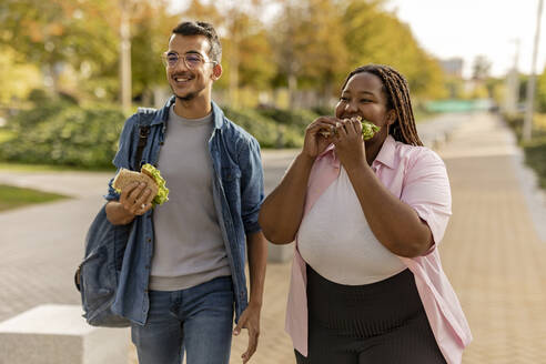 Happy young couple eating sandwich and walking on footpath - JCCMF08080