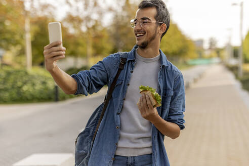 Happy young man taking selfie through smart phone at footpath - JCCMF08082