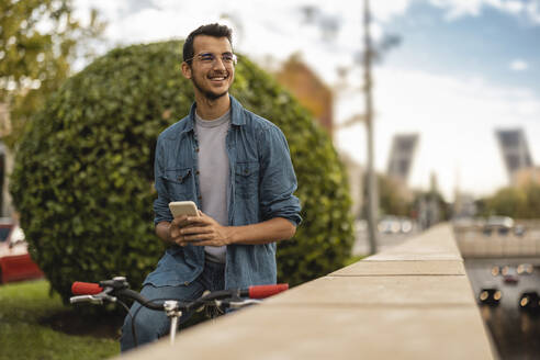 Glücklicher junger Mann mit Smartphone auf dem Fahrrad sitzend - JCCMF08120