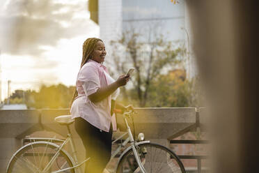 Glückliche junge Frau steht mit Fahrrad und benutzt ihr Smartphone - JCCMF08126