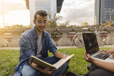Lächelnder junger Mann liest ein Buch mit einer Frau, die einen Laptop im Gras benutzt - JCCMF08128