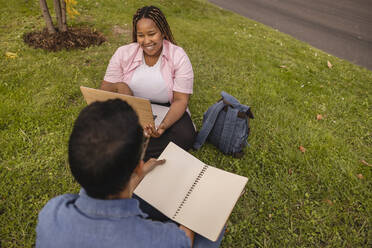 Happy young couple with laptop and book sitting on grass - JCCMF08130