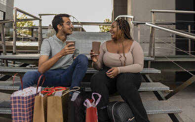 Young couple with shopping bags and coffee cups sitting on steps - JCCMF08143