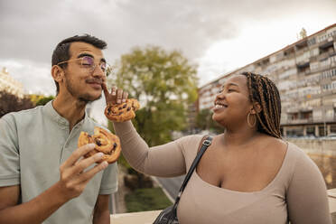 Happy young woman wiping boyfriend's face and standing with sweets under cloudy sky - JCCMF08149