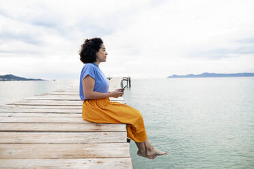 Frau sitzt mit Smartphone auf einem Steg am Meer - JOSEF14798