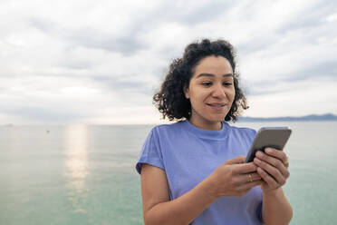 Lächelnde Frau mit Mobiltelefon vor dem Meer - JOSEF14804