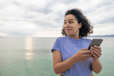 Lächelnde Frau steht mit Smartphone vor dem Meer - JOSEF14805