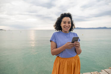 Lächelnde Frau mit Handy in der Hand vor dem Meer - JOSEF14807