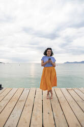 Lächelnde Frau auf dem Steg mit Smartphone vor dem Meer stehend - JOSEF14808