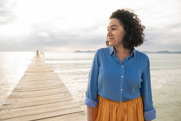 Lächelnde Frau mit lockigem Haar steht auf einem Steg am Meer - JOSEF14827