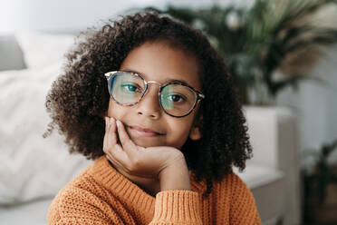 Smiling girl wearing eyeglasses sitting with hand on chin at home - EBBF07159