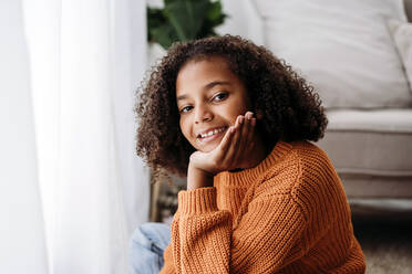 Happy cute girl sitting with hand on chin at home - EBBF07163