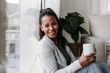 Smiling mature woman with coffee cup leaning on glass window at home - EBBF07172