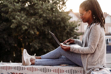 Lächelnde Frau mit Laptop auf der Terrasse bei Sonnenuntergang - EBBF07200