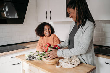 Lächelndes Mädchen mit Mutter, die Gemüse auf dem Tisch zu Hause hält - EBBF07215