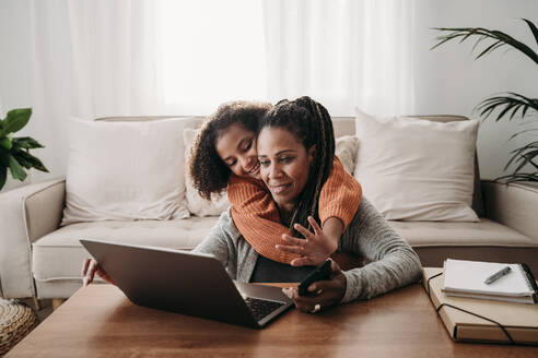 Lächelndes Mädchen umarmt Mutter mit Laptop für Videoanruf im Wohnzimmer zu Hause - EBBF07225