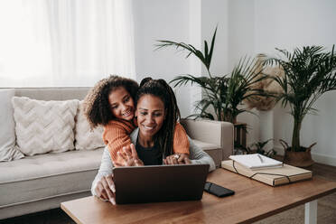 Happy girl with mother using laptop for video call in living room at home - EBBF07226