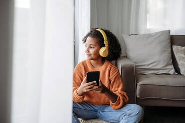 Smiling girl wearing wireless headphones sitting with smart phone at home - EBBF07239
