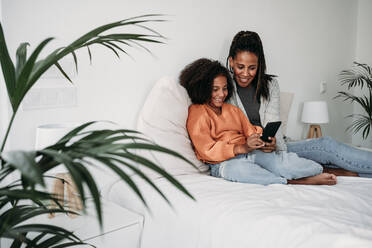 Happy mother and daughter using smart phone on bed at home - EBBF07244