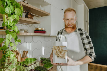 Smiling man holding oyster mushrooms in kitchen at home - YTF00325