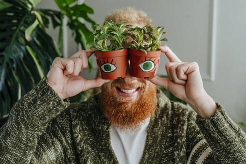 Happy man holding potted plants with eyes at home - YTF00343