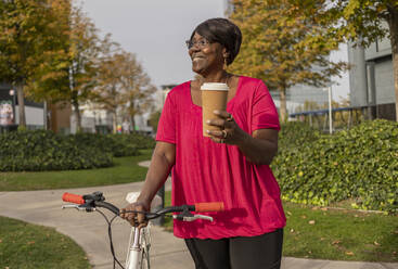 Lächelnde Frau mit Einwegbecher auf dem Fahrrad im Park - JCCMF08198