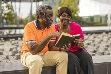 Senior couple reading book sitting on wall - JCCMF08216