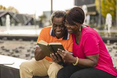 Smiling senior couple sharing book sitting on wall - JCCMF08218