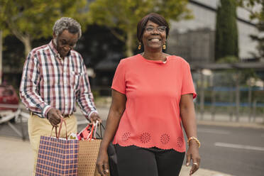 Lächelnde Frau geht mit einem Mann, der Einkaufstaschen im Hintergrund hält - JCCMF08230