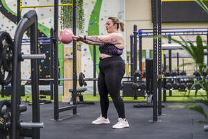 Junge übergewichtige Frau trainiert mit einer Kettlebell im Fitnessstudio - SNF01614