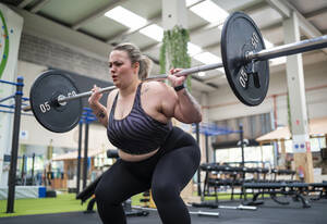 Übergewichtige Frau macht Kniebeugen mit Langhantel im Fitnessstudio - SNF01624